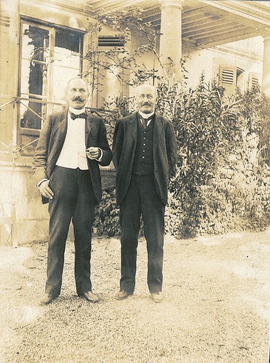 Photograph of two men dressed in suits standing next to each other in front of a house.