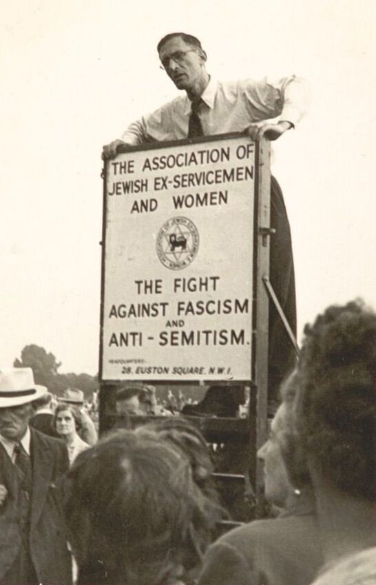 Black and white photograph of a man standing on top of a podium behind a sign which reads, 'The Association of Jewish Ex-Servicemen and Women' 'The Fight Against Fascism and Anti-Semitism'. People are gathered around the man looking up at him.