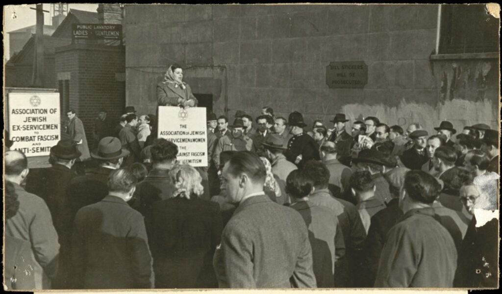 Black and white photograph of people gathered around a woman speaking. The woman is holding a sign. another sign in the background which reads, 'Association of Jewish Ex-Servicemen to Combat Fascism and Anti-Semitism'.
