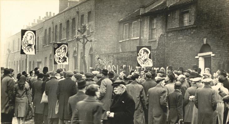 Black and white photograph of a crowd of people in a street gathered around a man speaking. Three signs held up with caricatures of Adolf Hitler, Oswald Mosley and Benito Mussolini drawn on them.