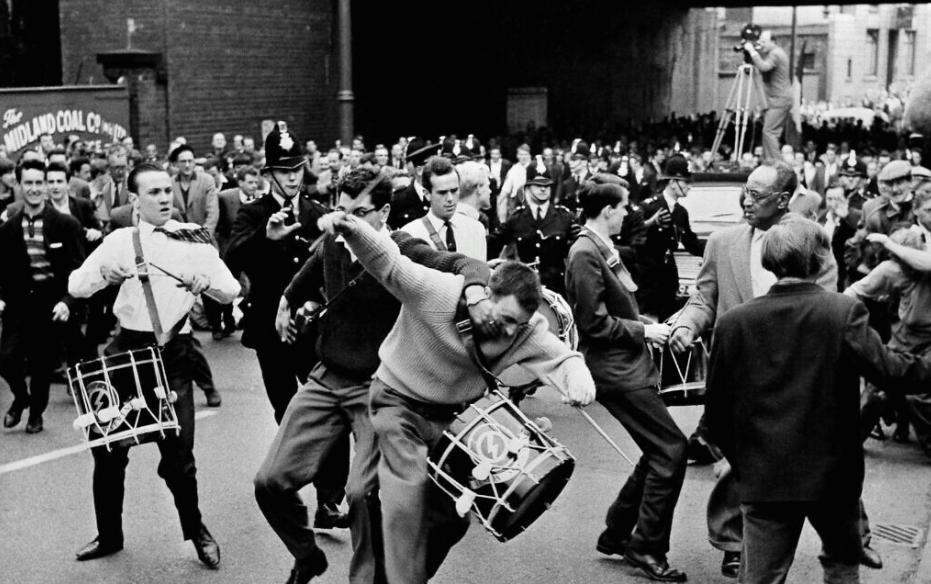 Three men carrying drums with the BUF logo on them being chased and attacked by other men. A crowd of people and police officers in the background.