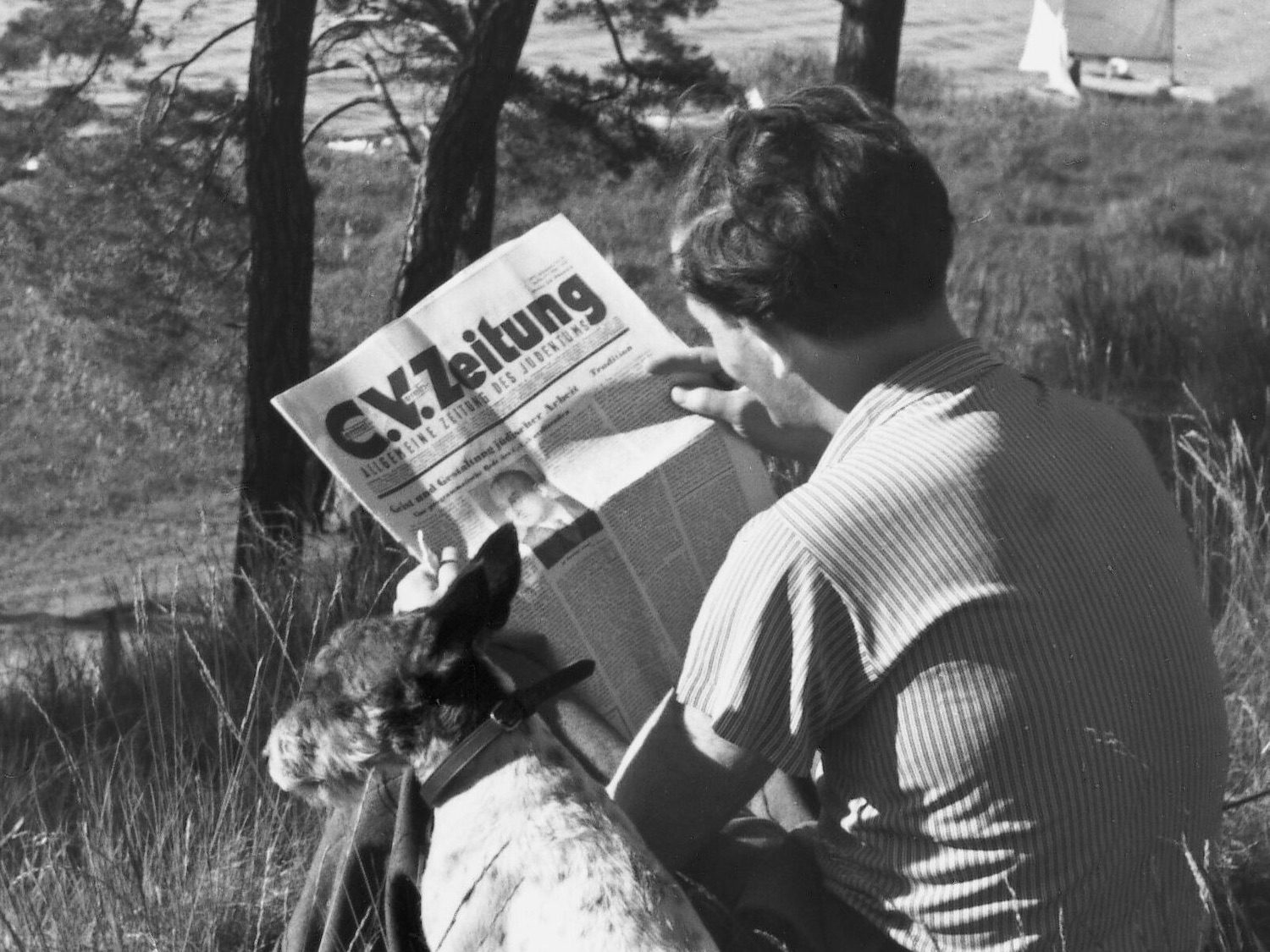 Man reading a newspaper whilst sitting in a field with a dog sitting beside him.